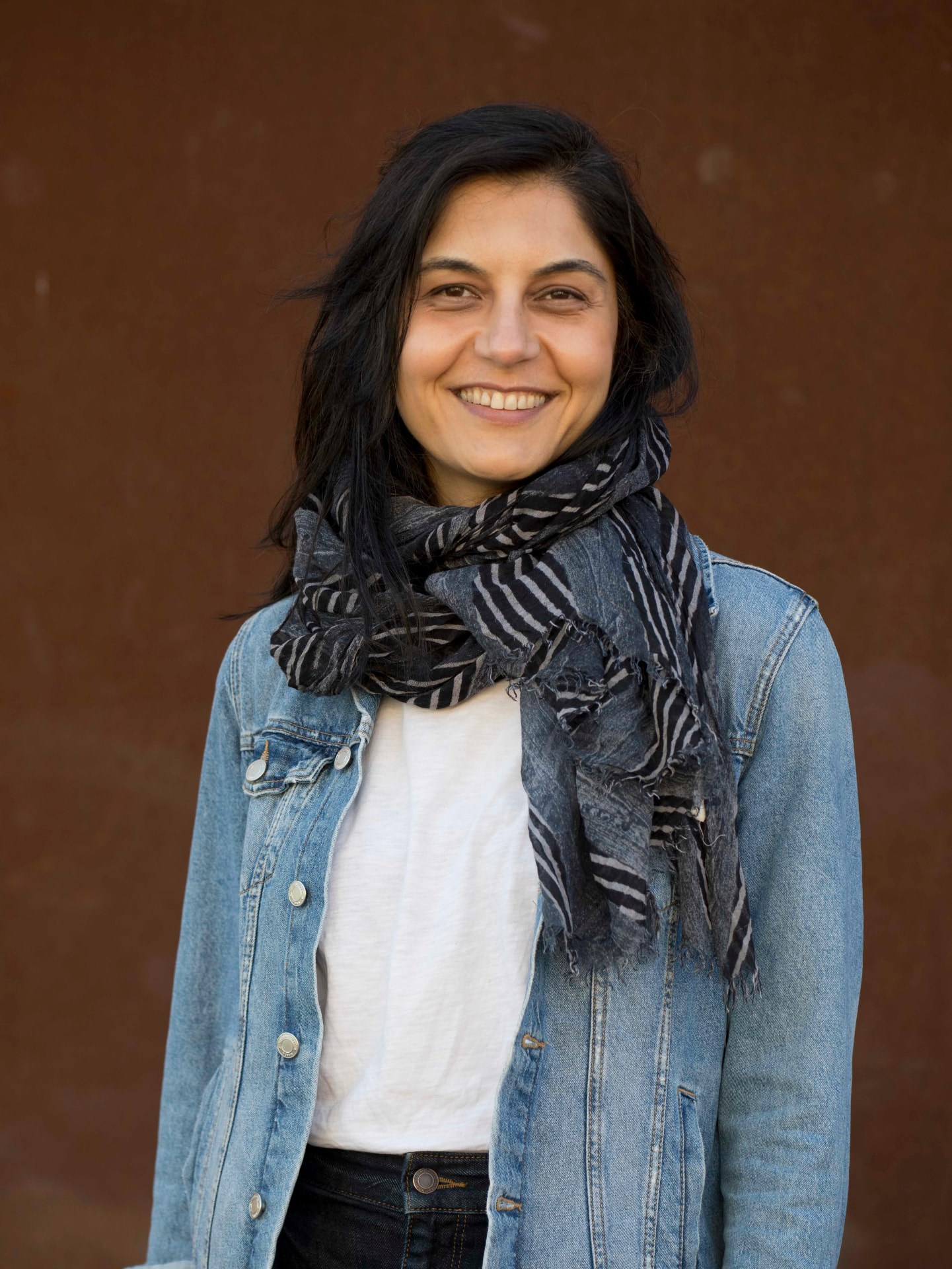 Portrait of Gunes Kocabag, smiling, wearing a denim jacket and striped scarf against a warm brown background